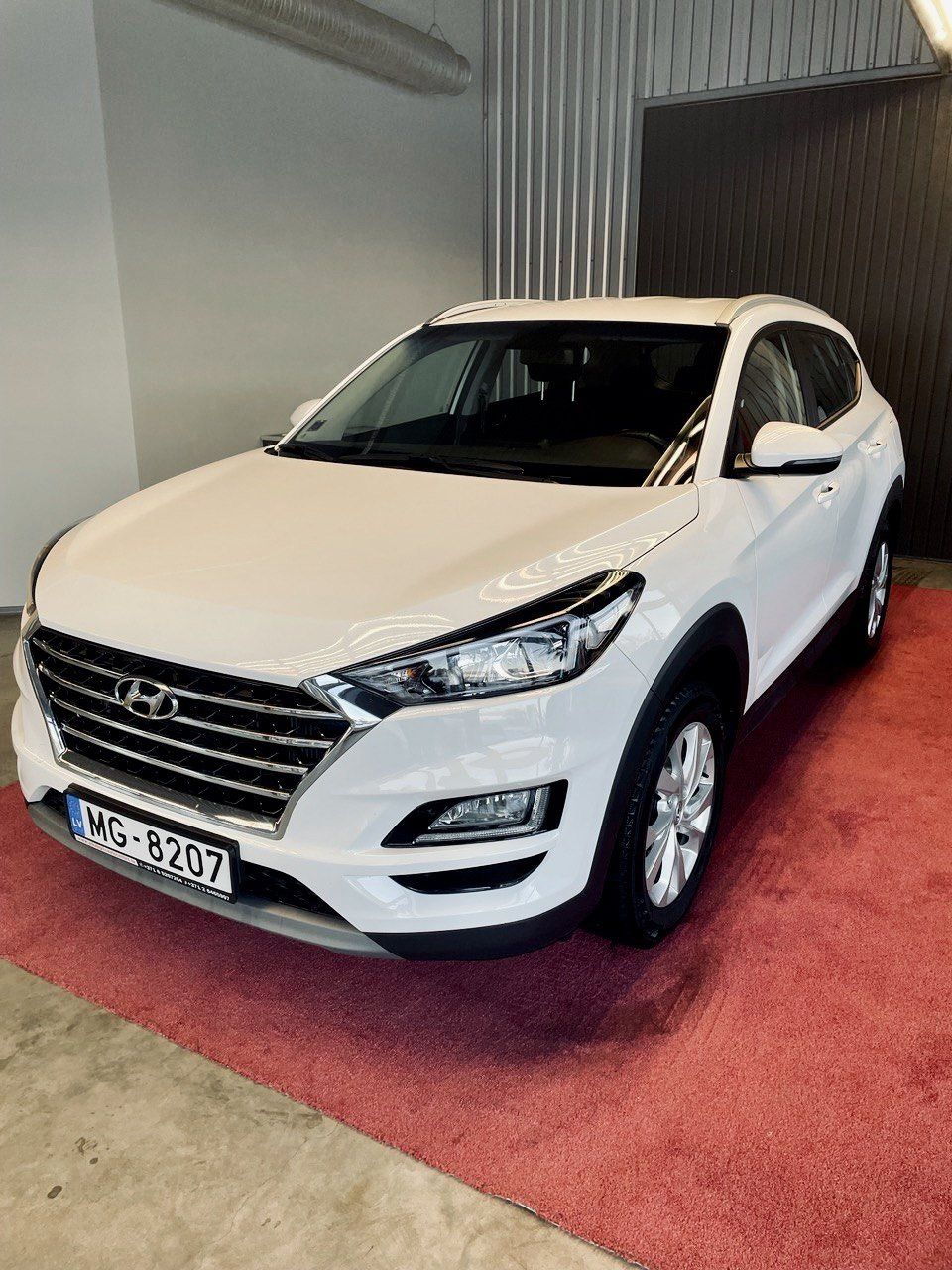 Balts hyundai tucson ir novietots garāžā uz sarkana paklāja.