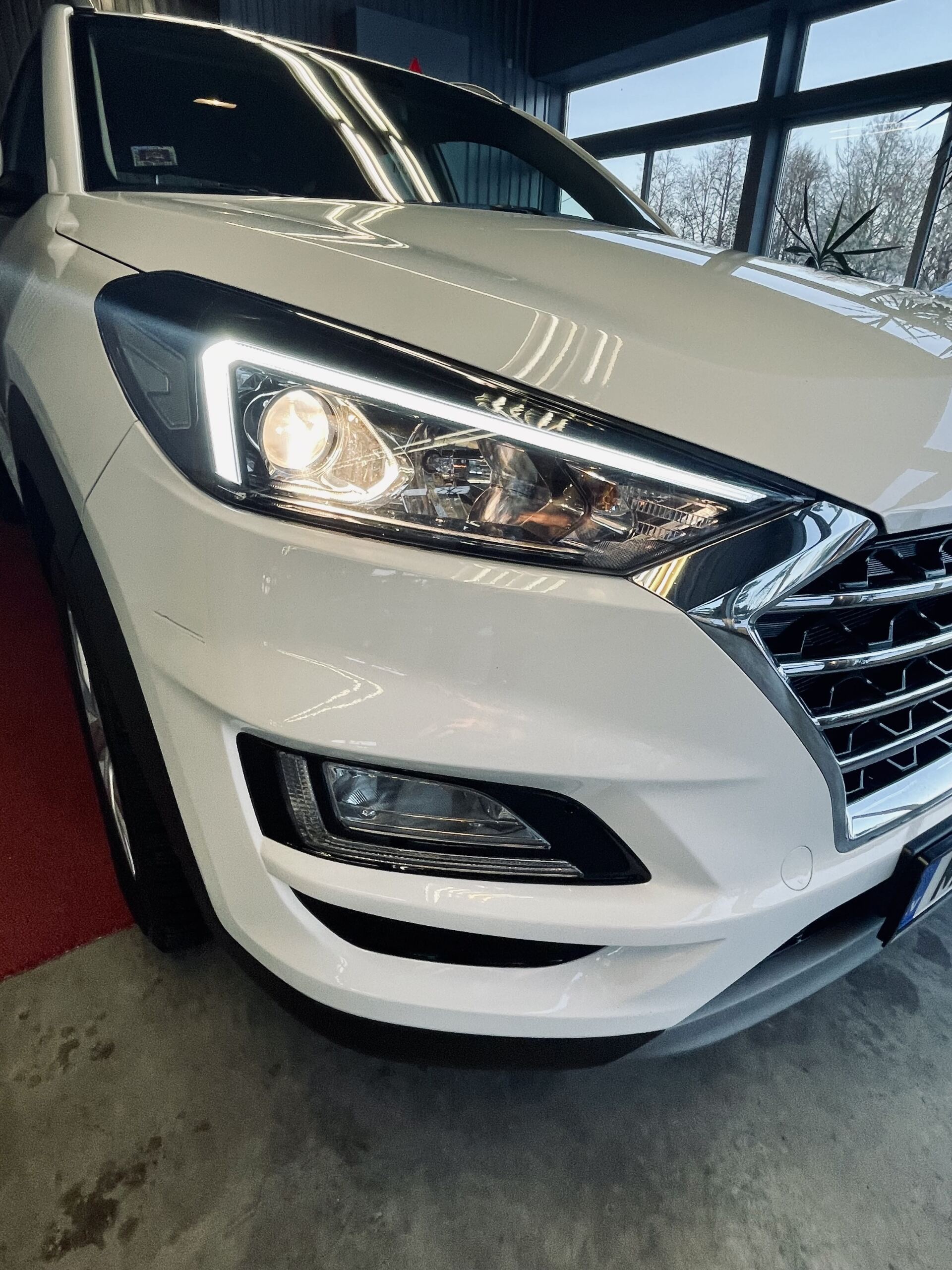 Balta automašīna ir novietota garāžā ar ieslēgtiem lukturiem.