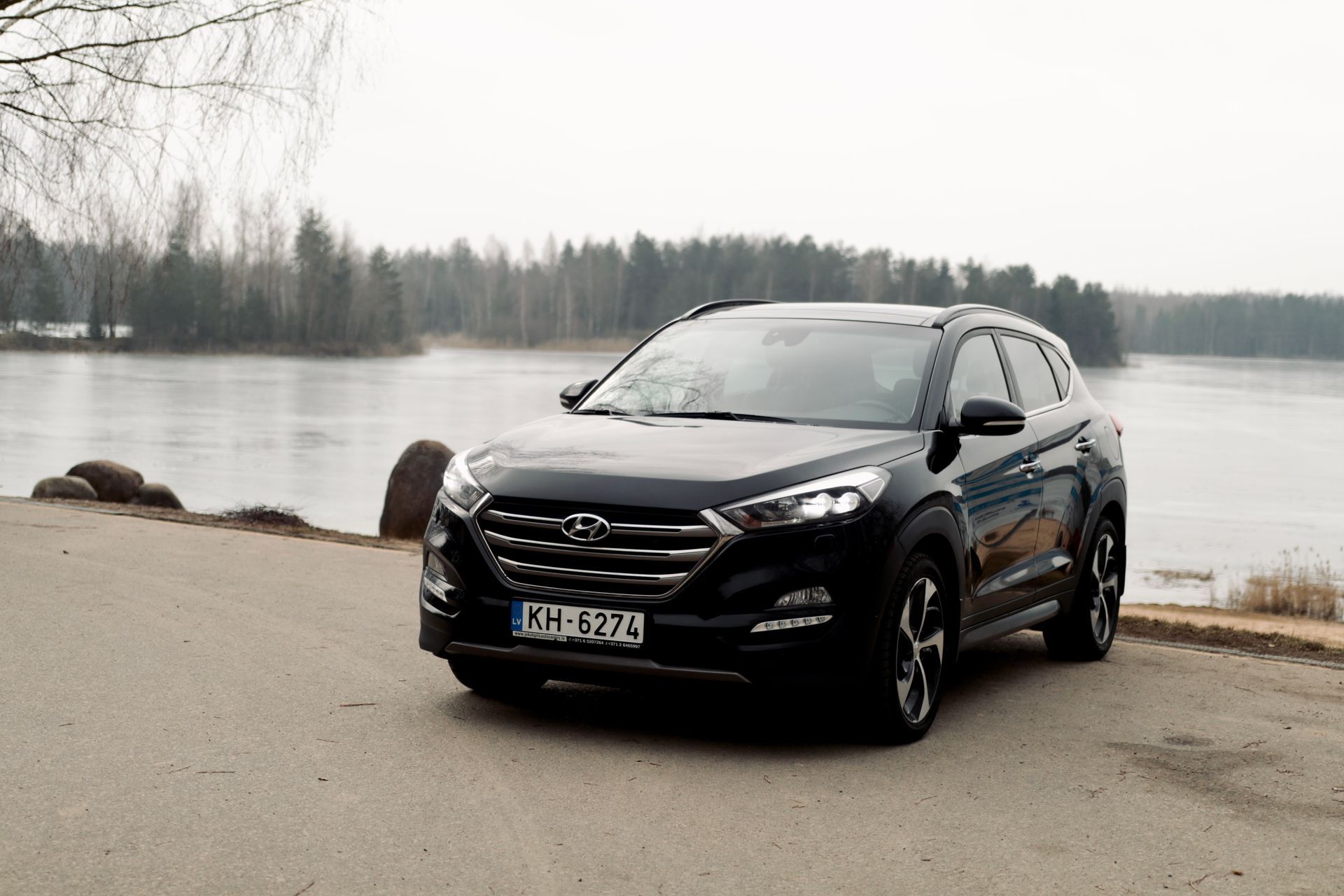 Ceļa malā pie ezera stāv melna mašīna.