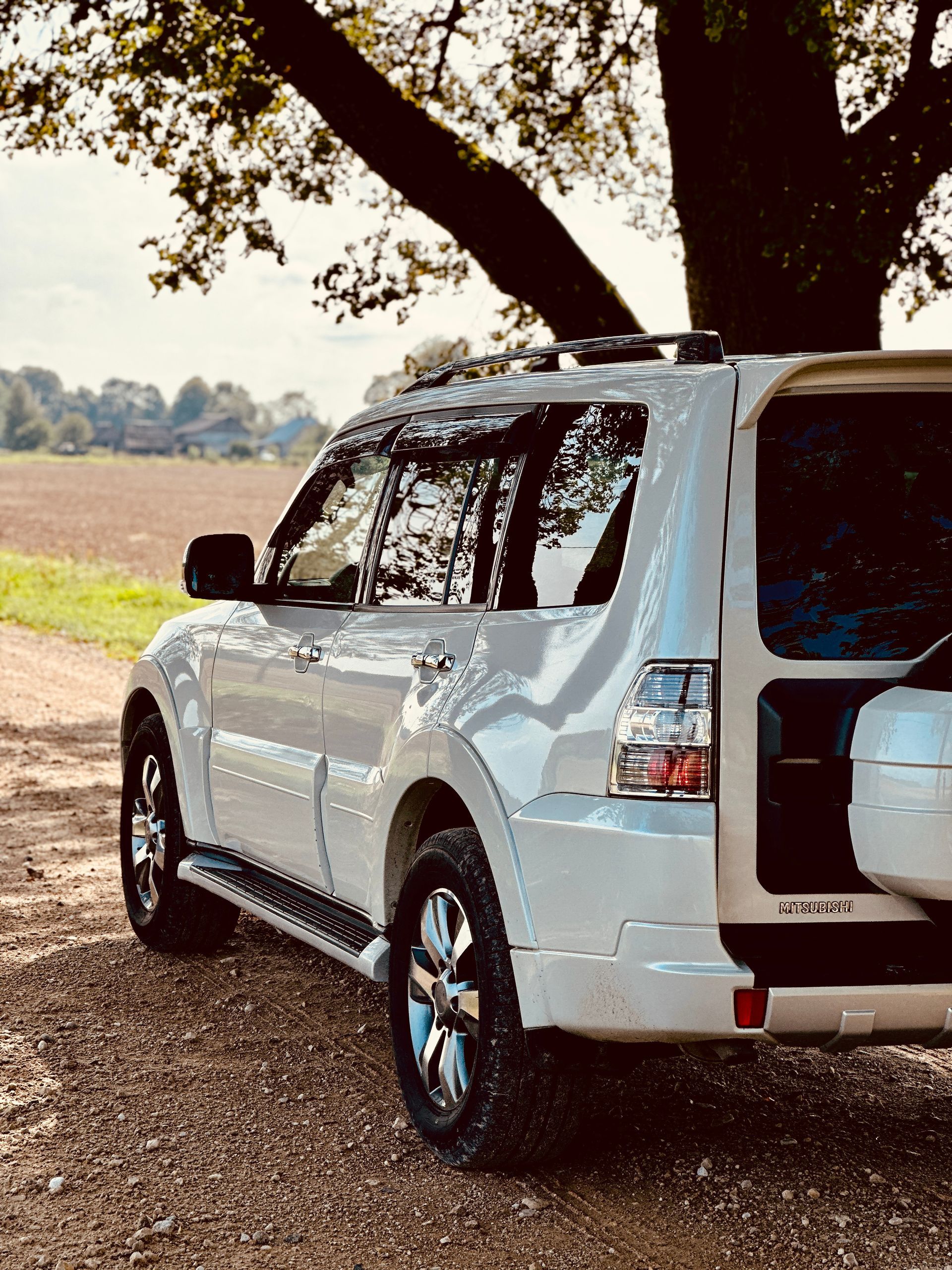 Balts apvidus auto stāv zem koka uz zemes ceļa.
