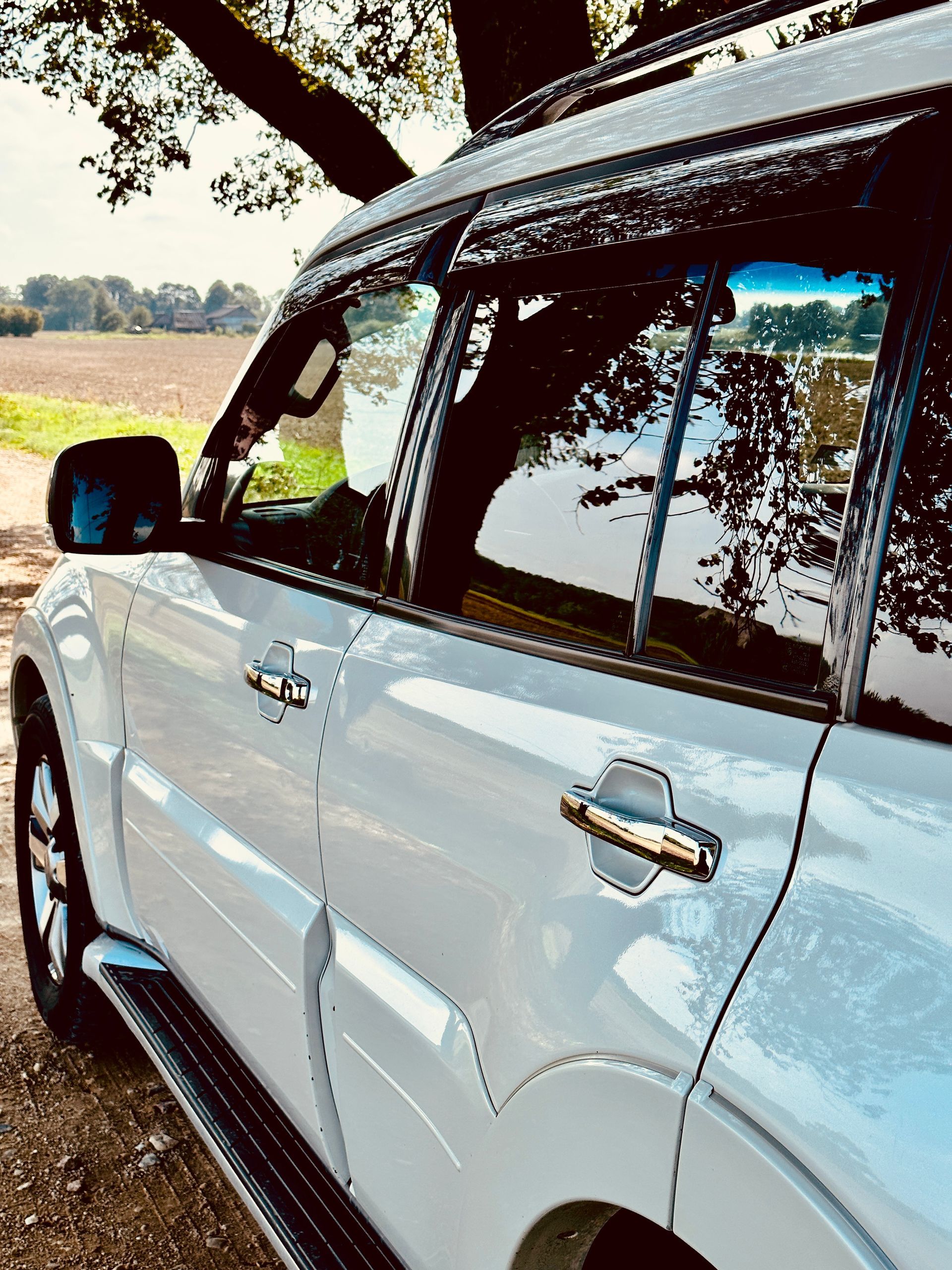 Balts apvidus auto stāv zem koka uz zemes ceļa.