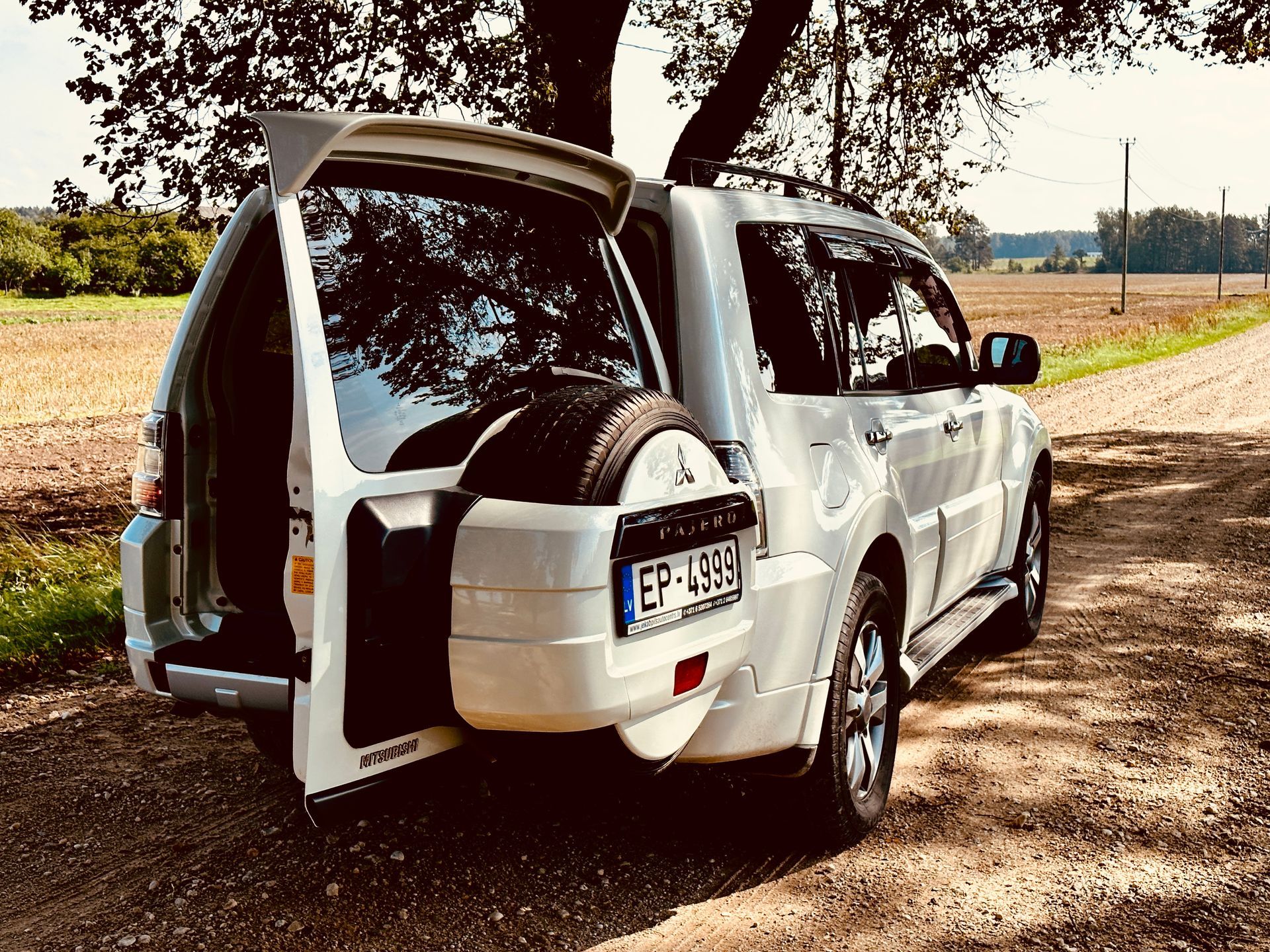 Balts apvidus auto ar atvērtu bagāžnieku stāv uz zemes ceļa.