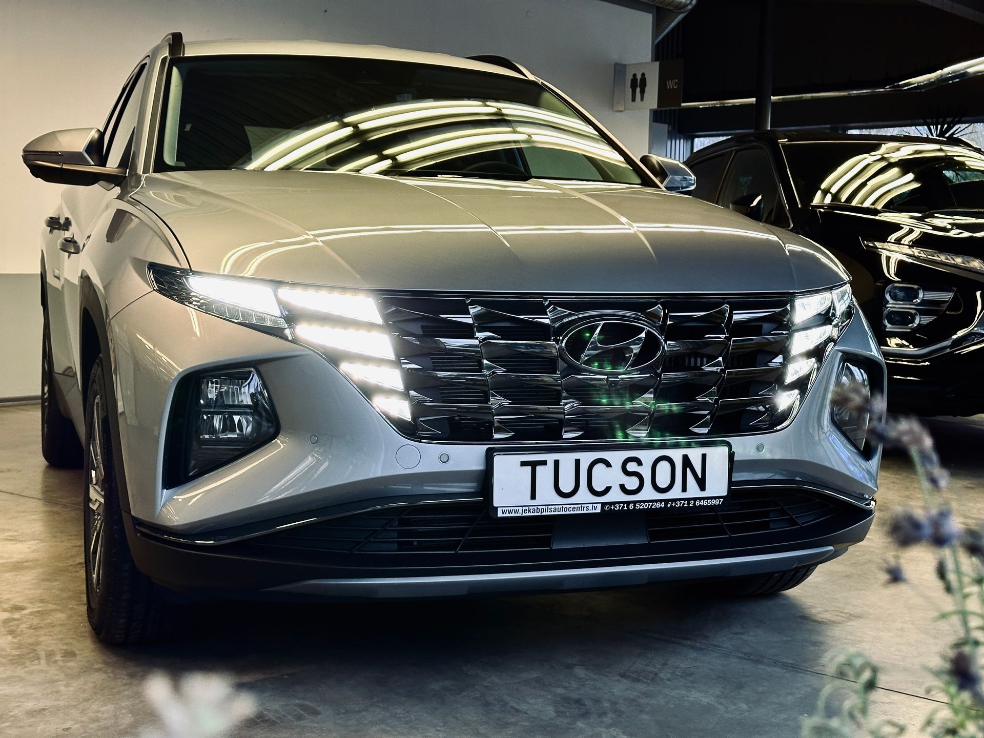 Hyundai tucson stāv salonā pie melnas mašīnas.