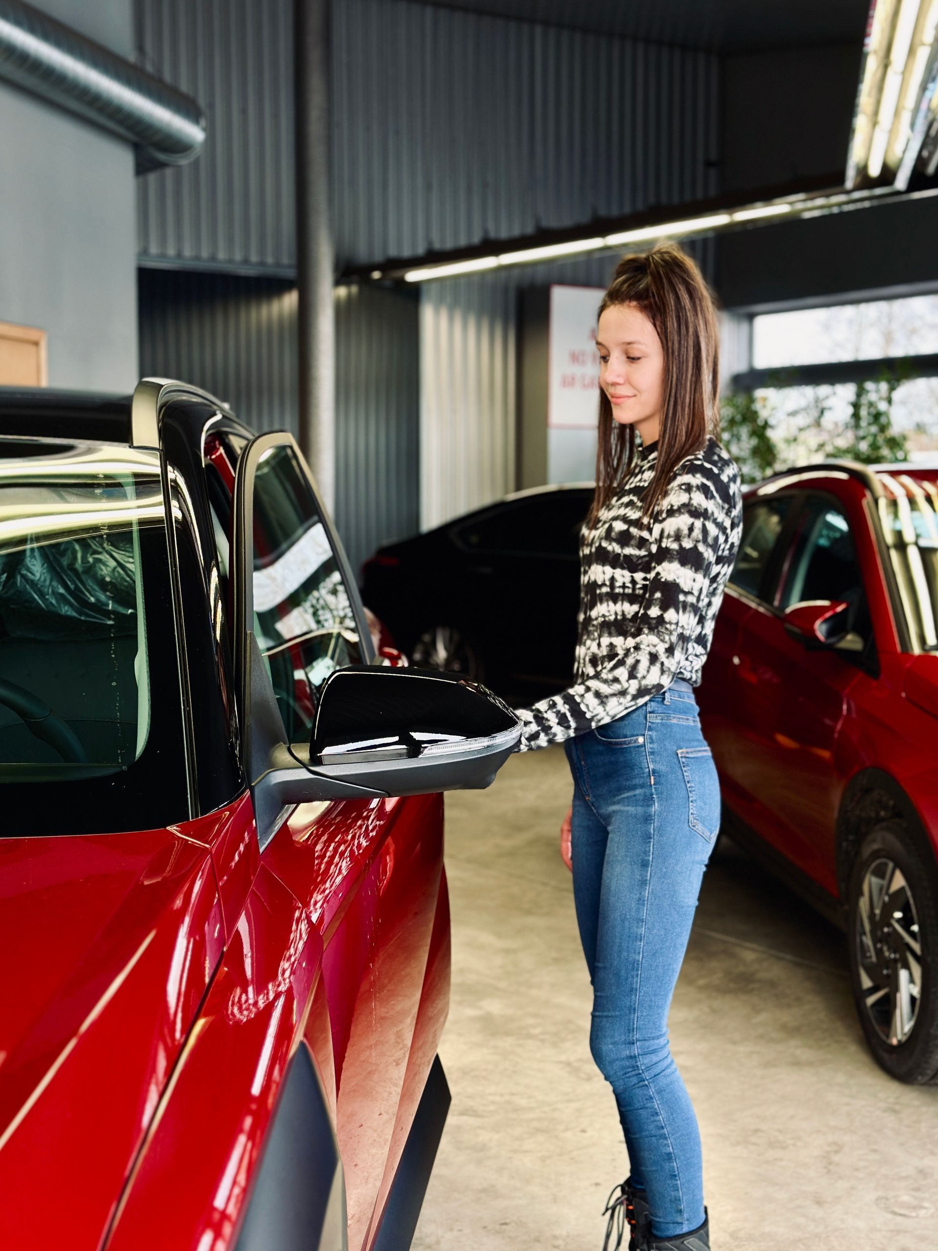 Sieviete stāv garāžā pie sarkanas mašīnas.