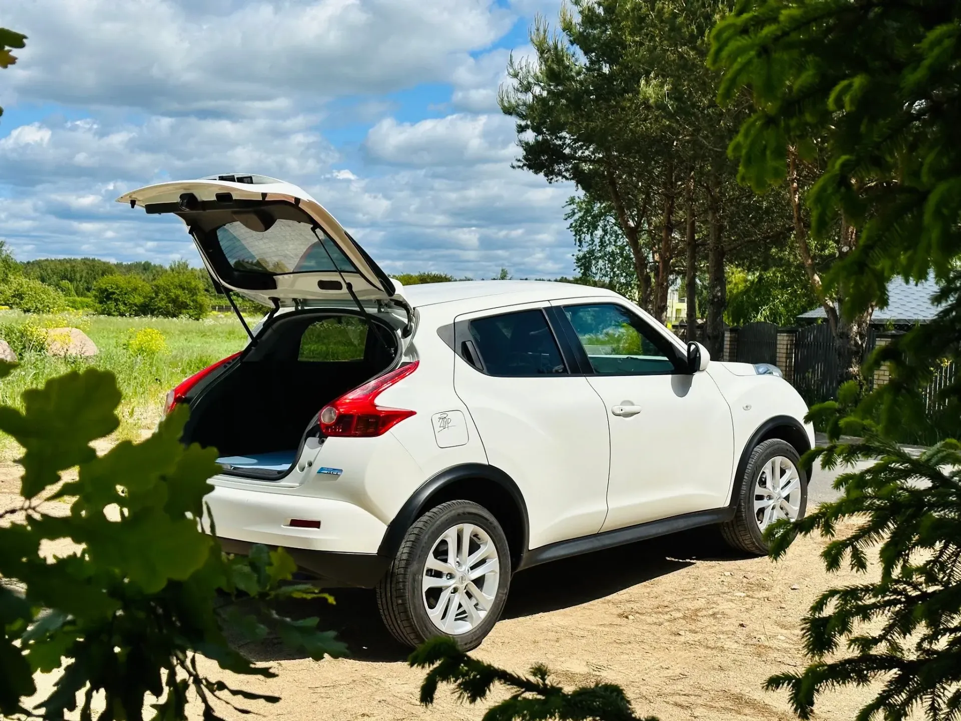 Uz zemes ceļa stāv balta mašīna ar atvērtu bagāžnieku.