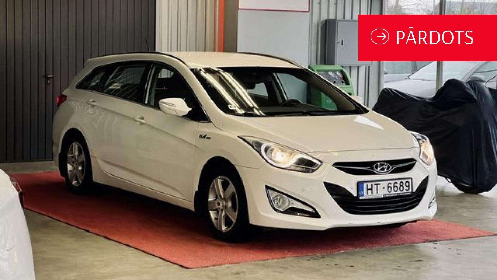 Balta mašīna stāv garāžā pie motocikla.