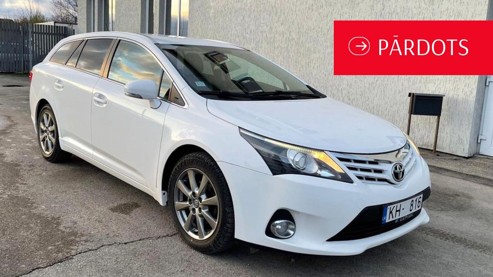Balta toyota avensis ir novietota pie ēkas.