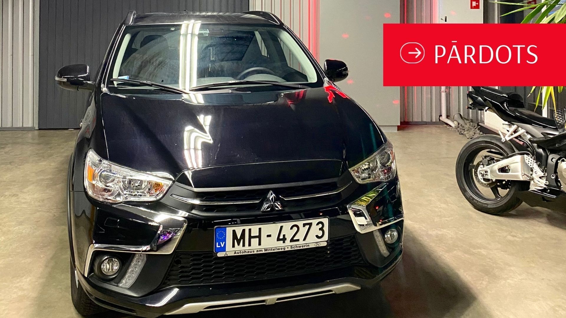 Melna mašīna stāv garāžā pie motocikla.