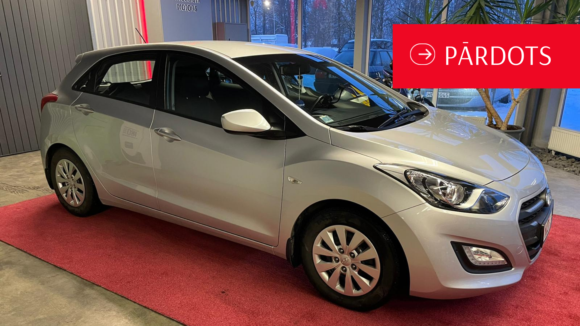 Sudraba automašīna ir novietota uz sarkanā paklāja izstāžu zālē.