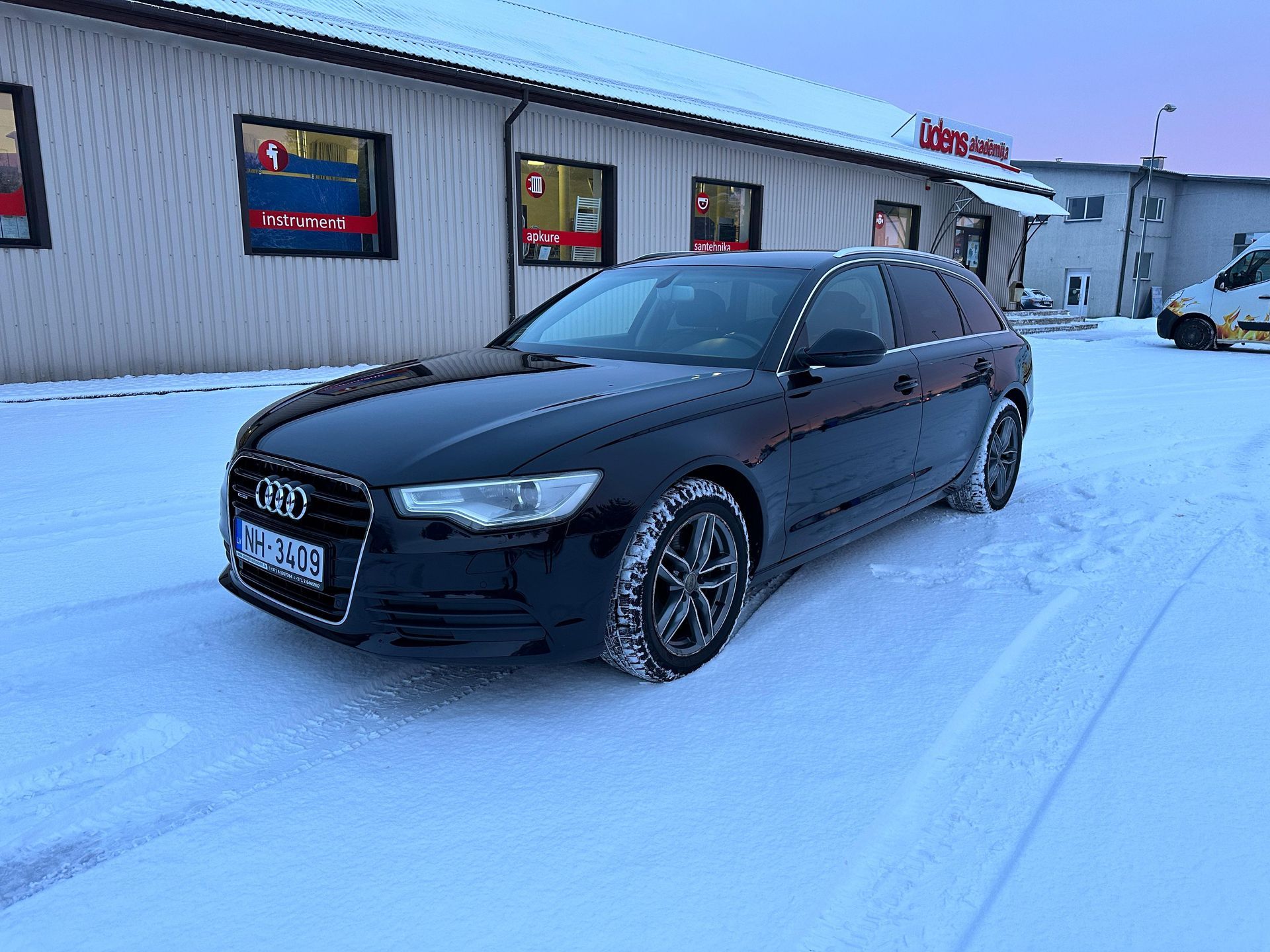 Melns audi a6 stāv sniegā pie ēkas.