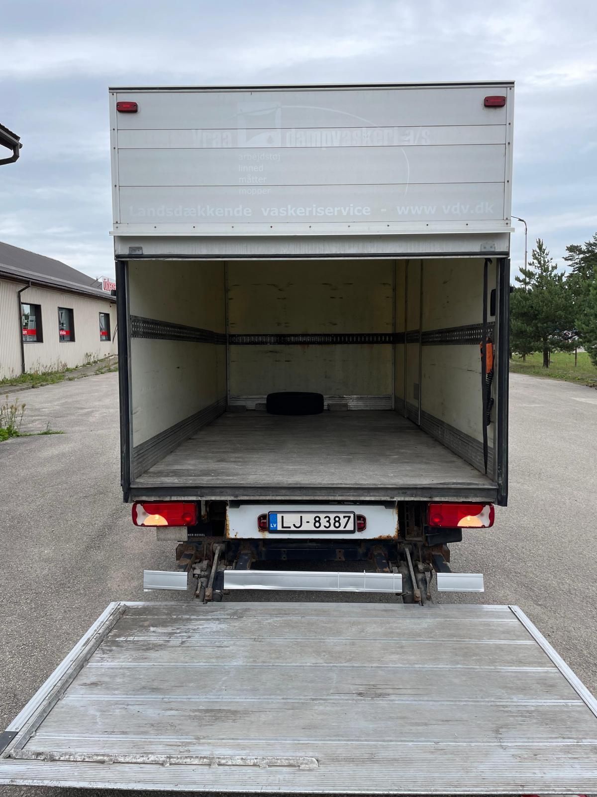 Ceļa malā stāv kravas automašīna ar atvērtām aizmugurējām durvīm.