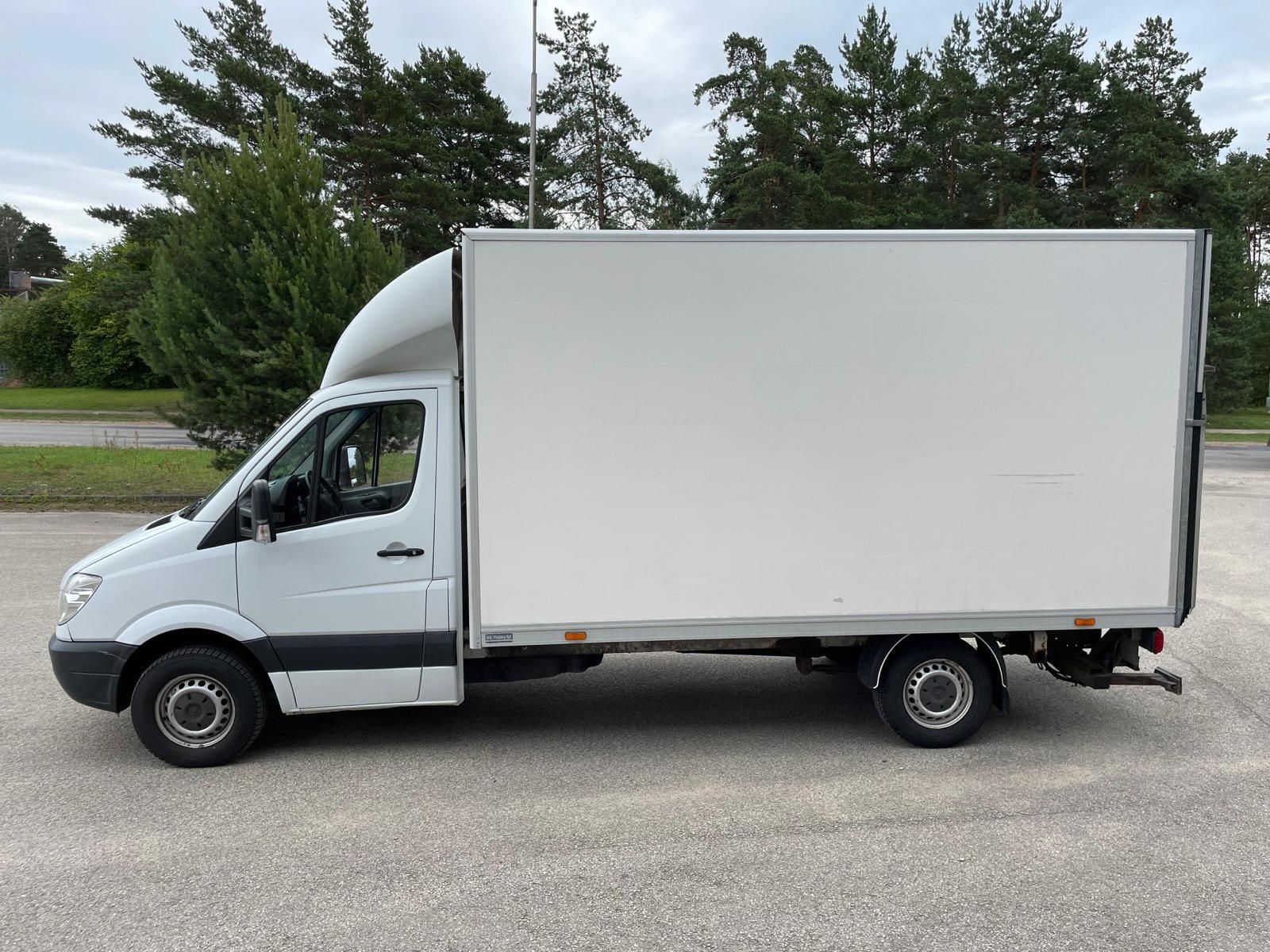 Balts furgons stāv stāvvietā ar kokiem fonā.