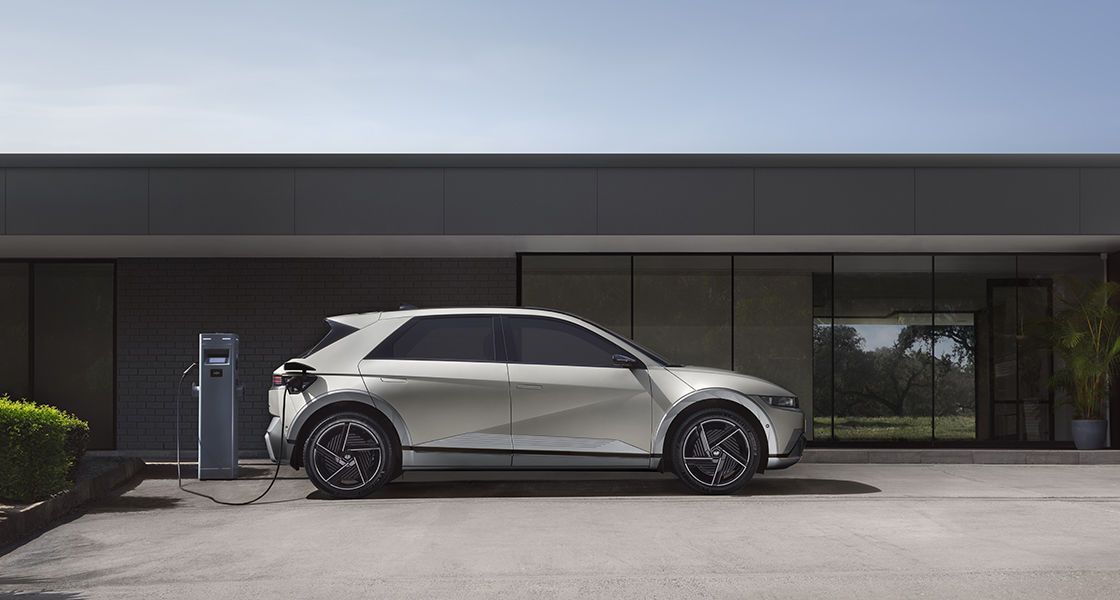 Pie mājas tiek uzlādēts balts elektromobilis.