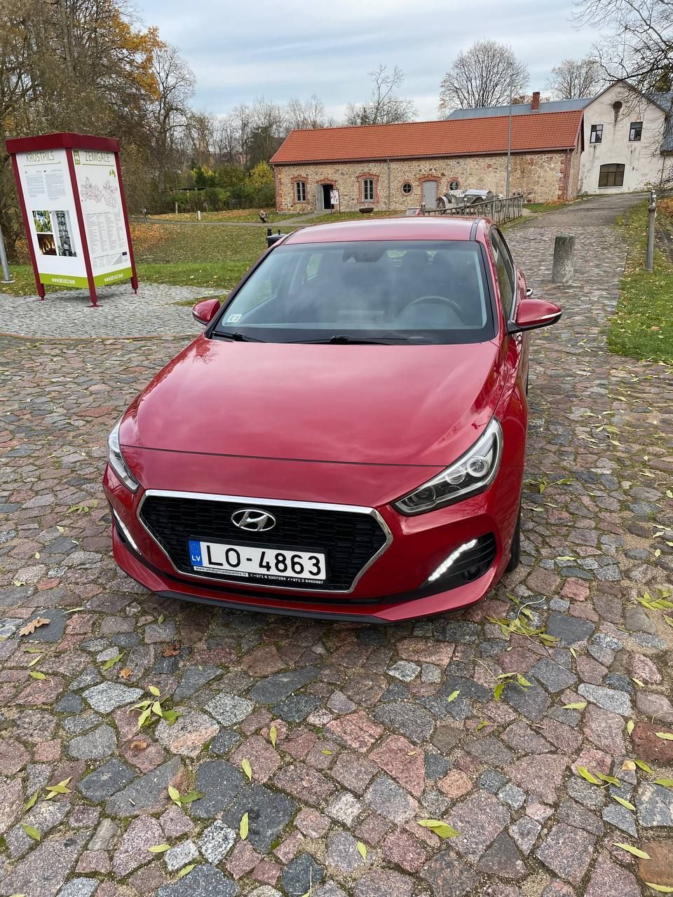 Sarkana automašīna ir novietota uz bruģakmens ceļa ēkas priekšā.