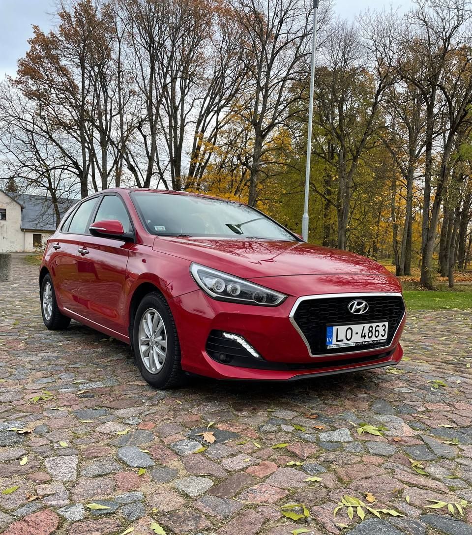 Uz bruģētas ielas koku priekšā stāv sarkana mašīna.