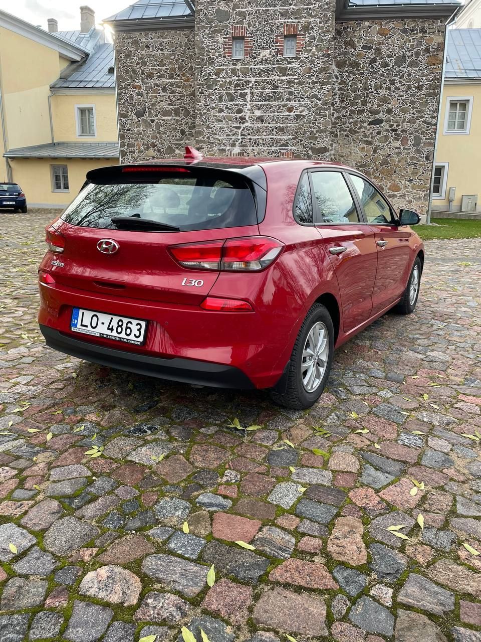 Sarkana automašīna ir novietota uz bruģakmens ielas iepretim mūra ēkai.