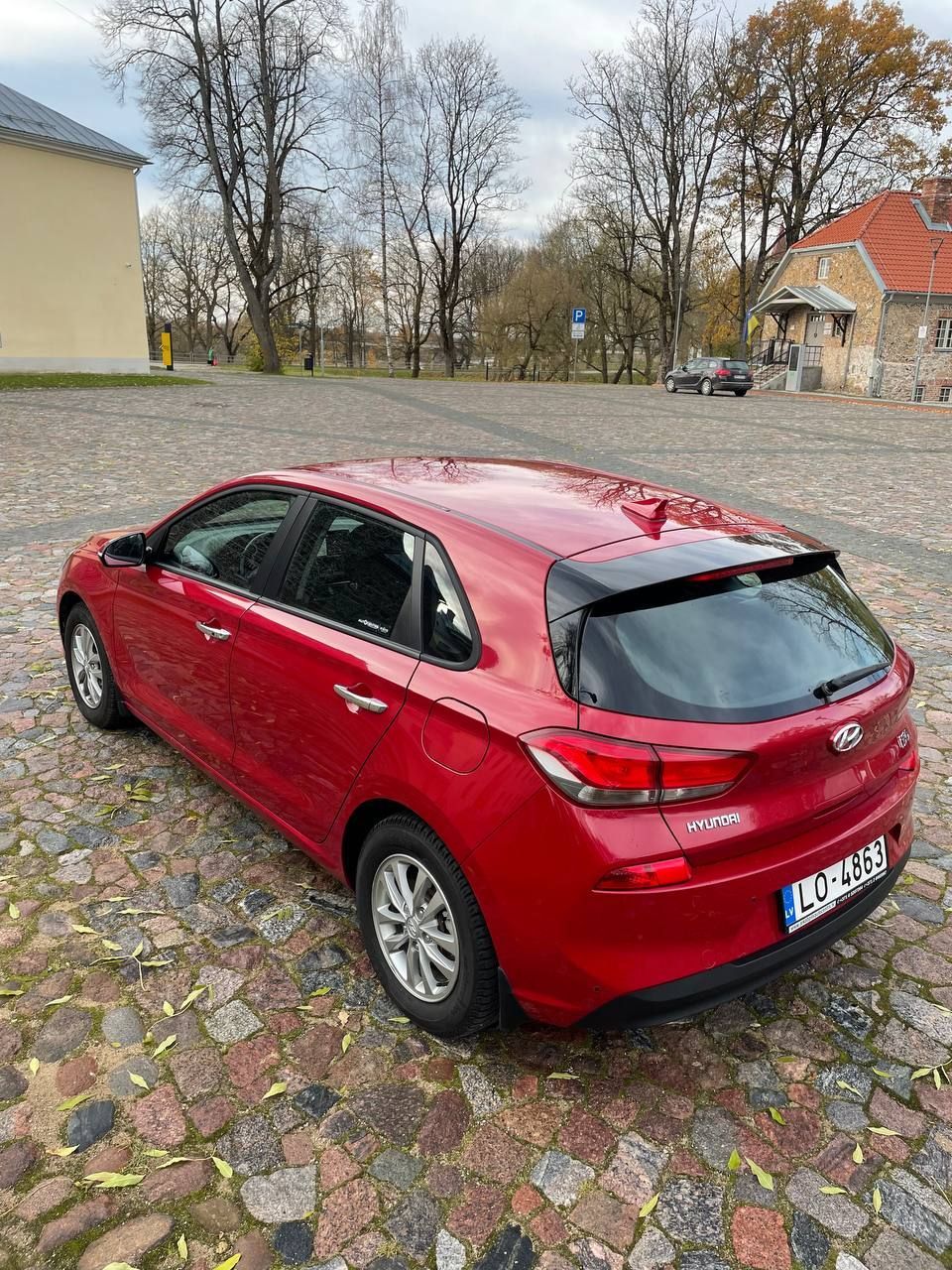 Sarkana automašīna ir novietota uz bruģakmens ielas ēkas priekšā.