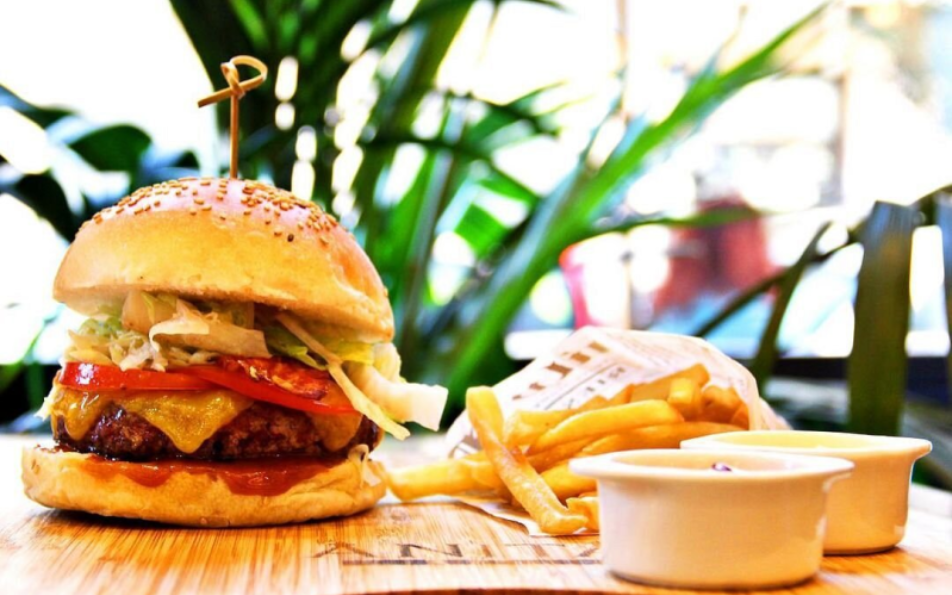 Hamburger con patatine fritte e salse su un tagliere di legno; vegetazione sullo sfondo.