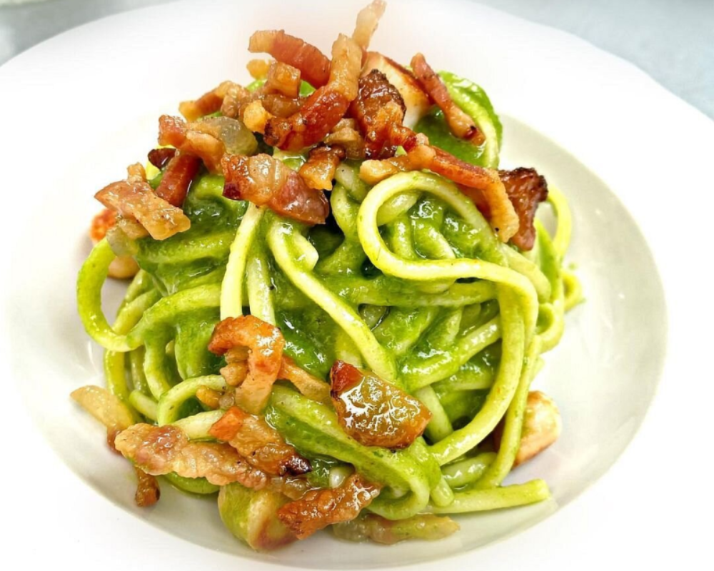 Piatto di pasta verde con pancetta sbriciolata sopra, servito in una ciotola bianca.