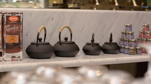Teiere nere con manici in legno su una mensola di marmo, con cioccolata e barattoli sullo sfondo.