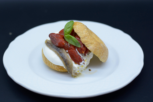 Panino con acciughe, peperoni rossi, mozzarella e basilico su un piatto bianco.