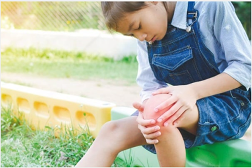 A little girl is sitting on a green bench holding her knee in pain.