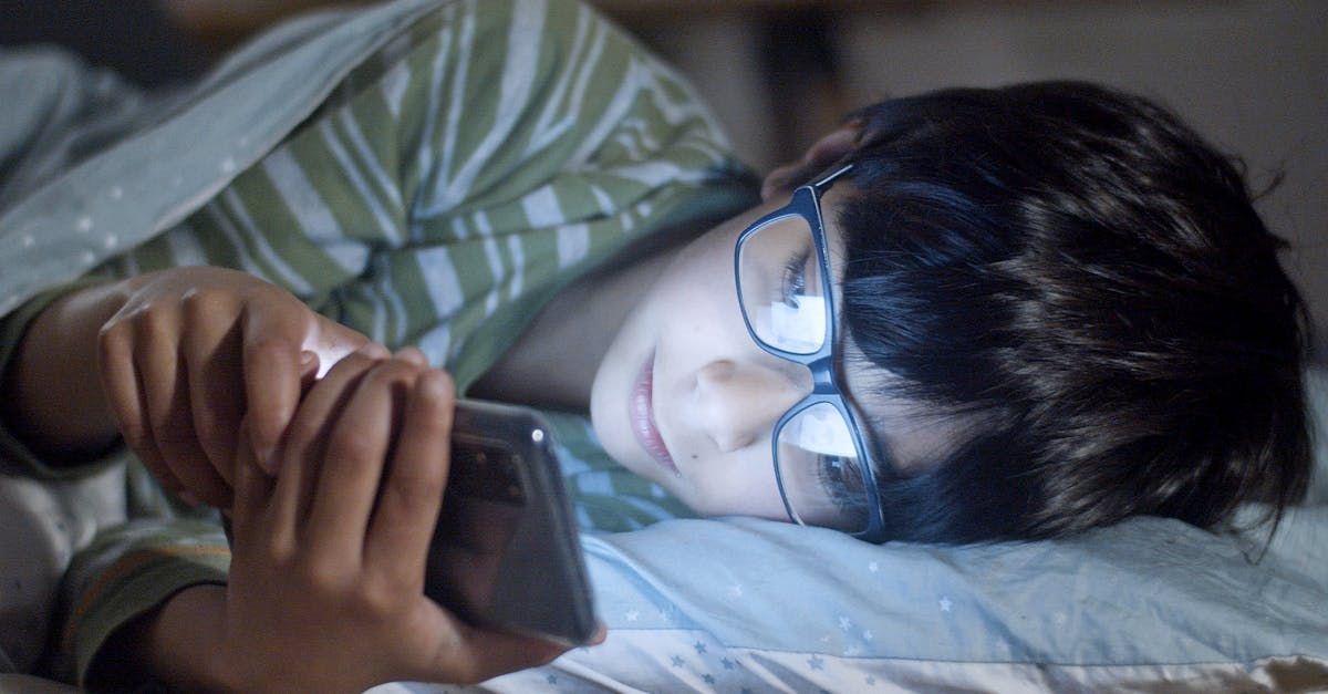 A young boy wearing glasses is laying in bed using a cell phone.