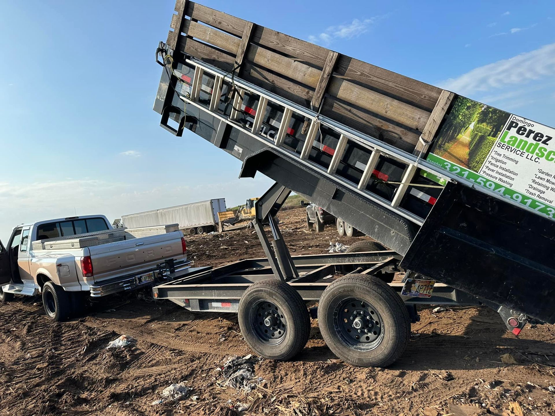 A dump truck is being towed by a trailer in a dirt field.
