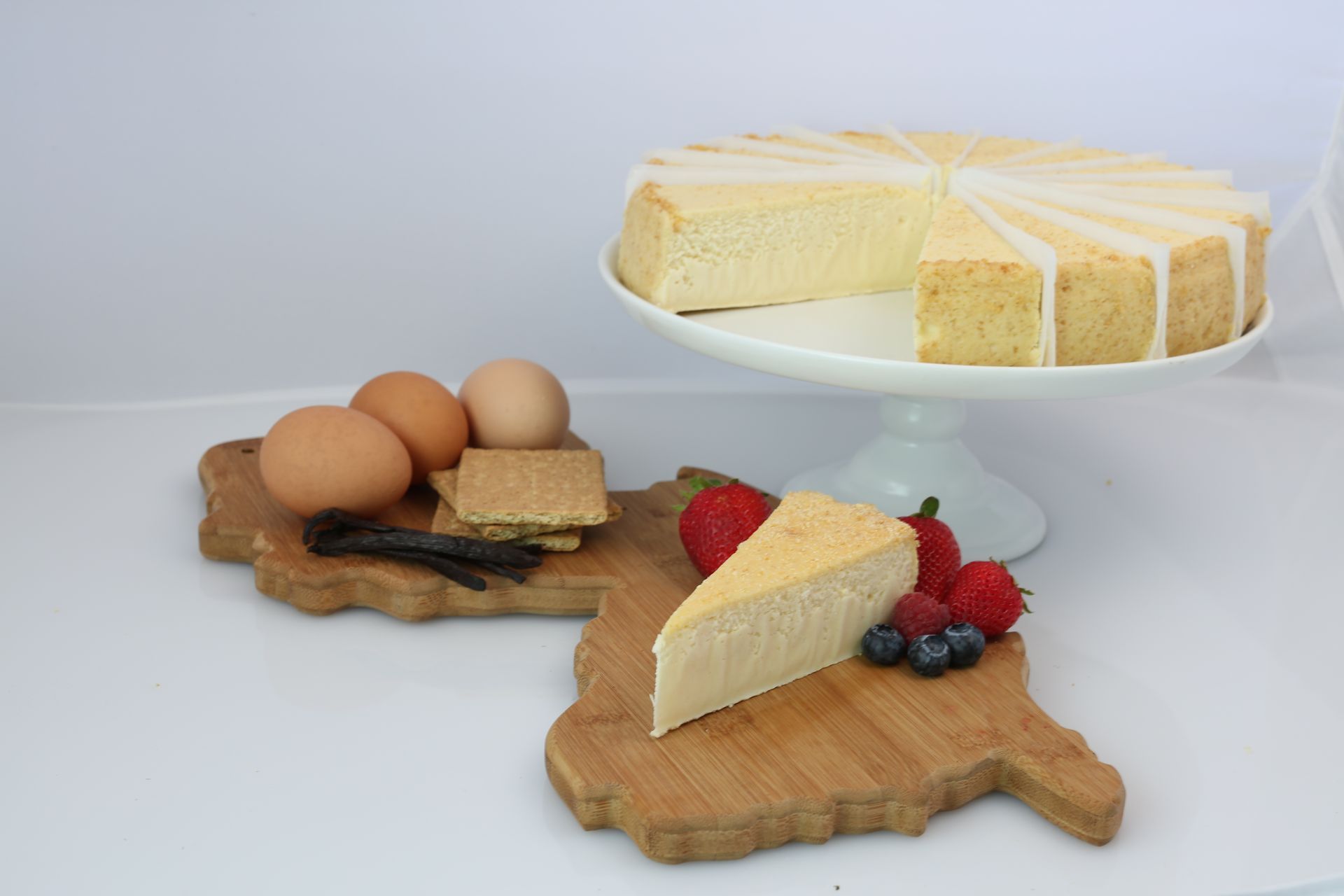A slice of cheesecake is on a wooden cutting board next to eggs and berries.