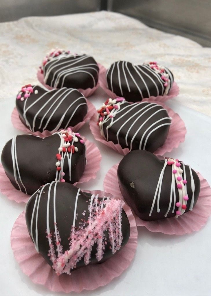 Heart-shaped chocolate treats with white drizzle and pink sprinkles, in pink paper cups.