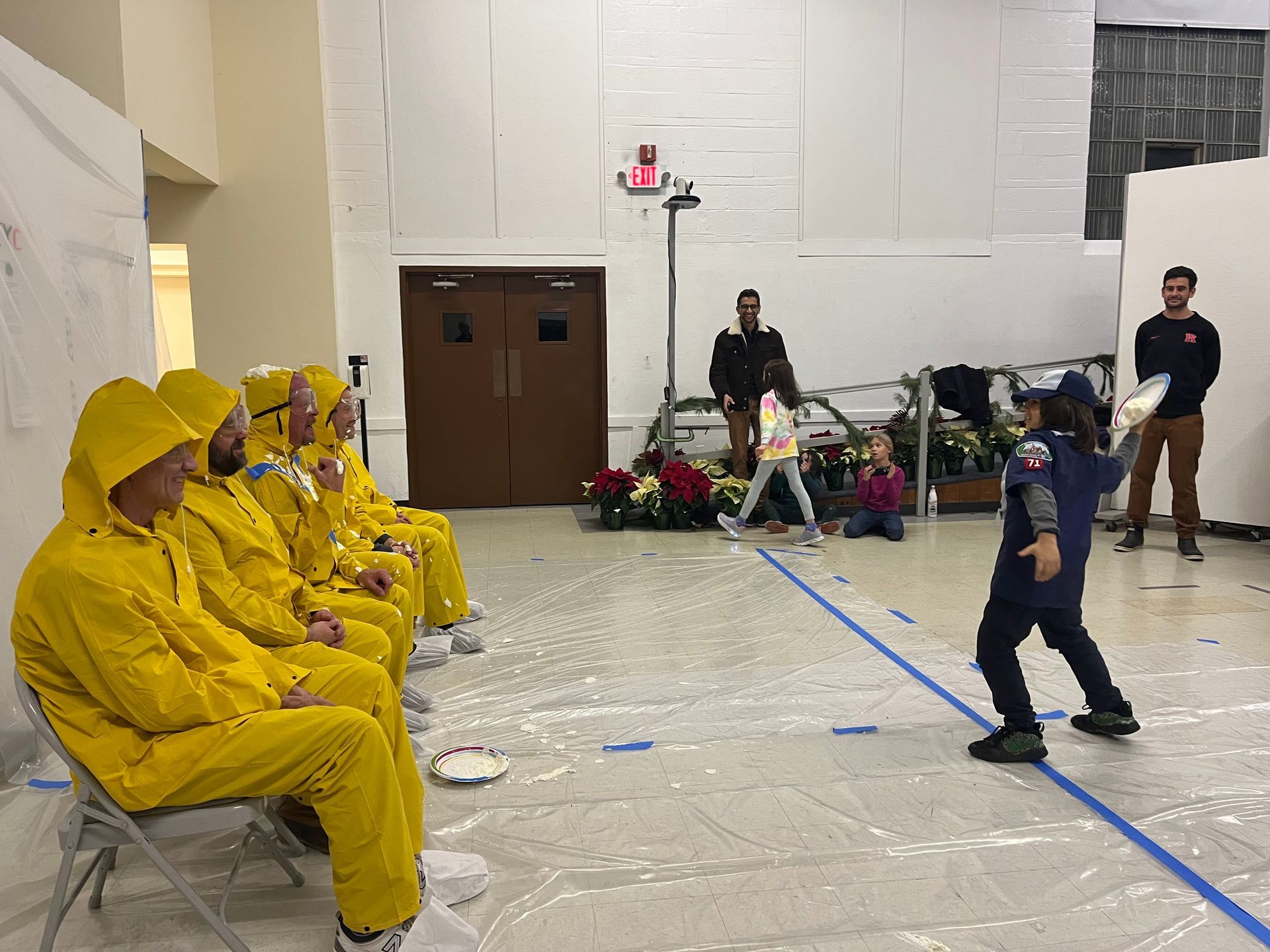 A child throws a pie at people in yellow suits seated on chairs, inside a hall.
