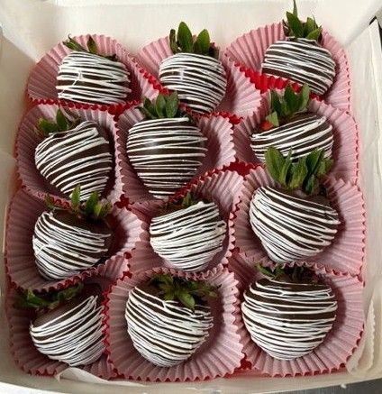 Chocolate-covered strawberries in individual pink paper cups inside a white box. Dark chocolate with white drizzle.