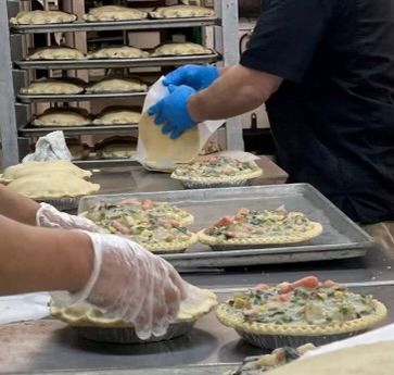 People preparing pies in a bakery; filling and crusting. Metal racks of pies in background.