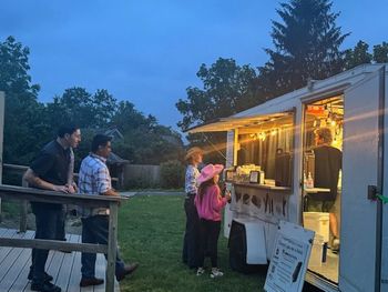 People ordering from a food truck at dusk; illuminated serving window.