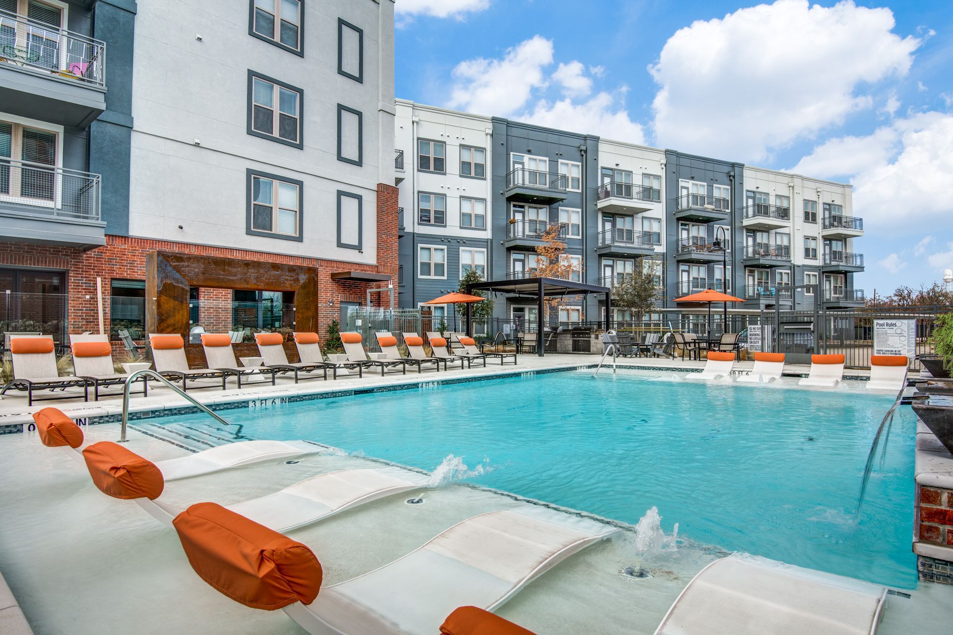 A large swimming pool surrounded by chairs and umbrellas in front of a building at Two99 Monroe offers apartments for rent in Roanoke, TX, near Downtown.