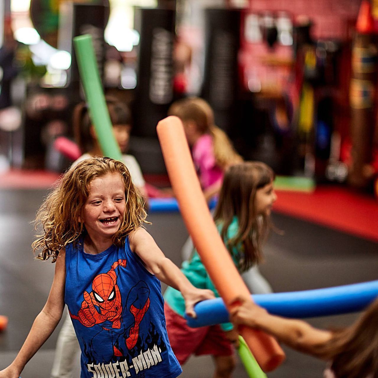 A little girl wearing a spiderman shirt is playing with foam sticks.