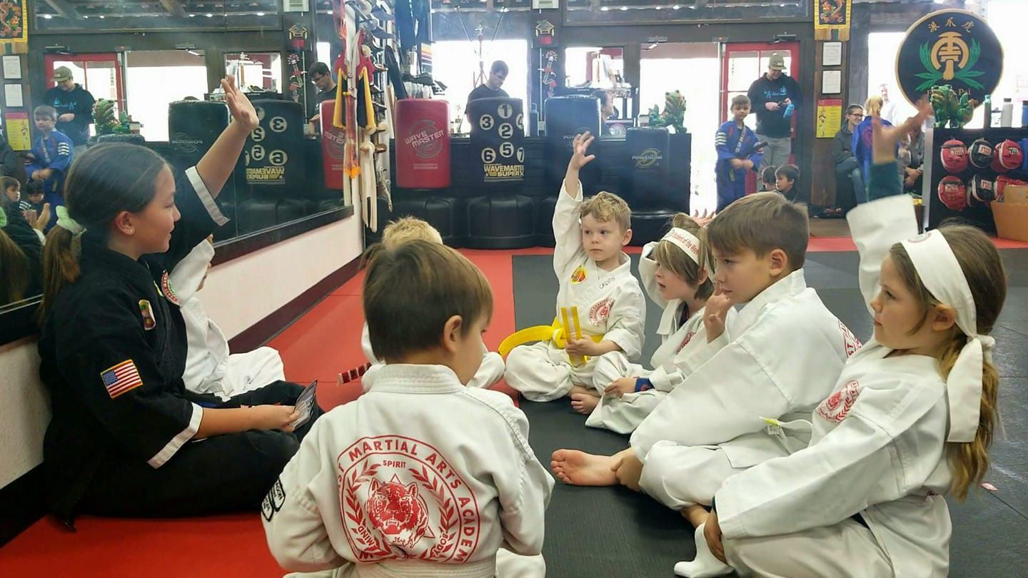 A group of children in karate uniforms are sitting on the floor in a gym.