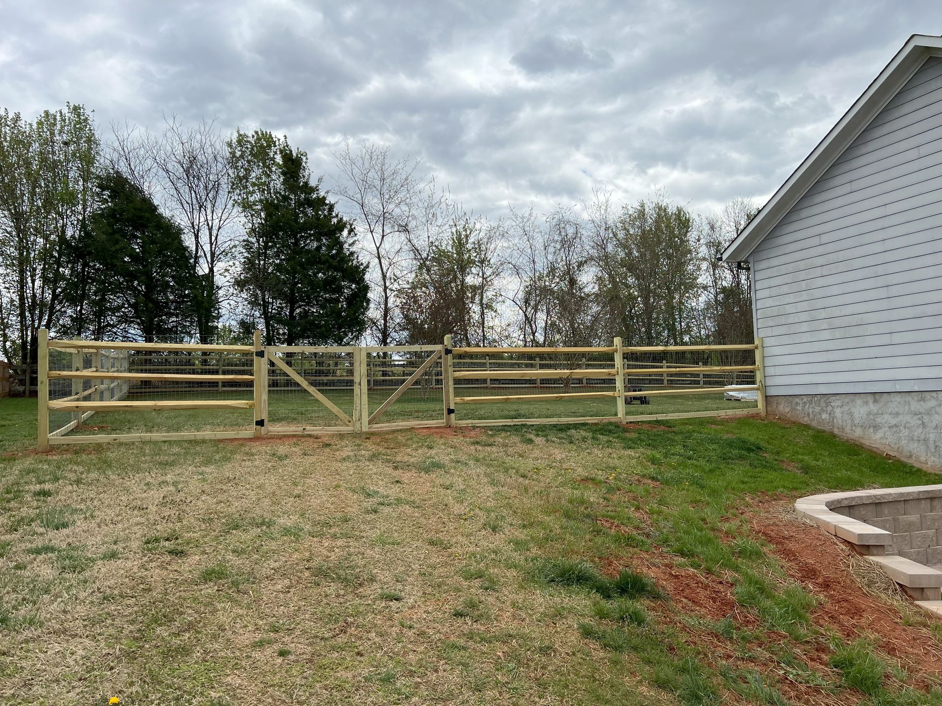 A wooden split-rail fence surrounds a grassy yard, with a house on the right and trees in the background under a cloudy sky.