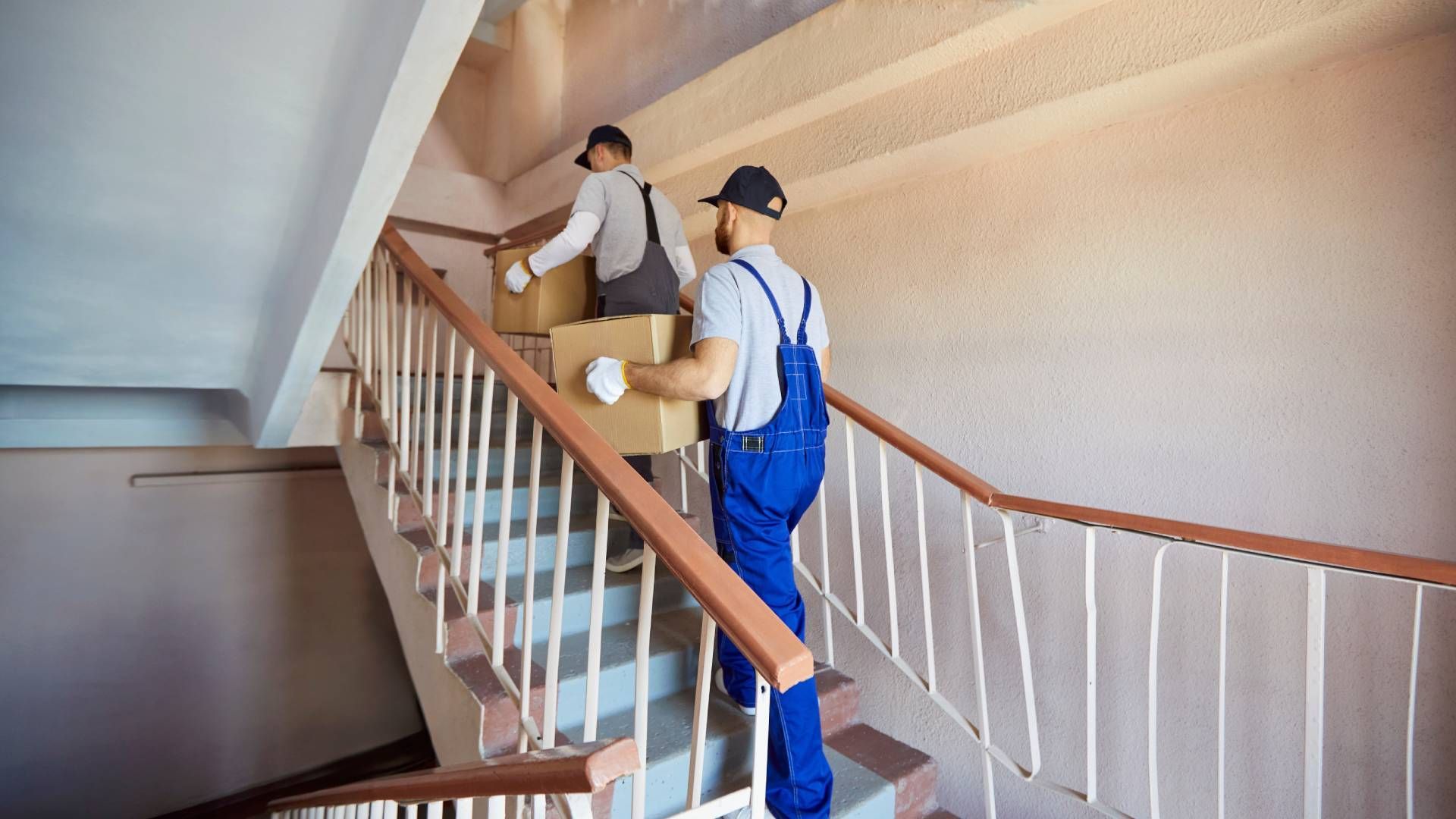Two movers in blue overalls and caps carrying boxes up a staircase.