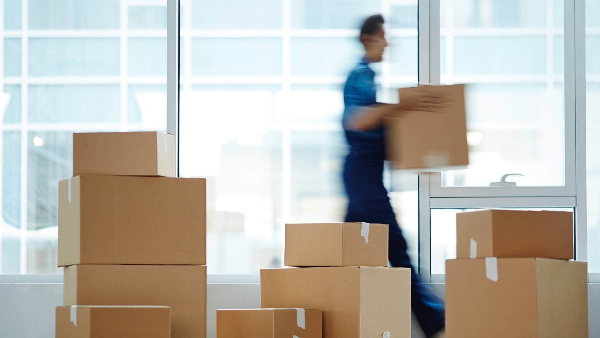 A person carrying a cardboard box in a room with stacked boxes and a large window.