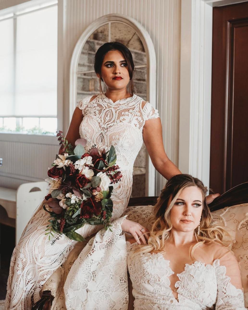 Two brides are posing for a picture while one is sitting on a couch holding a bouquet of flowers.