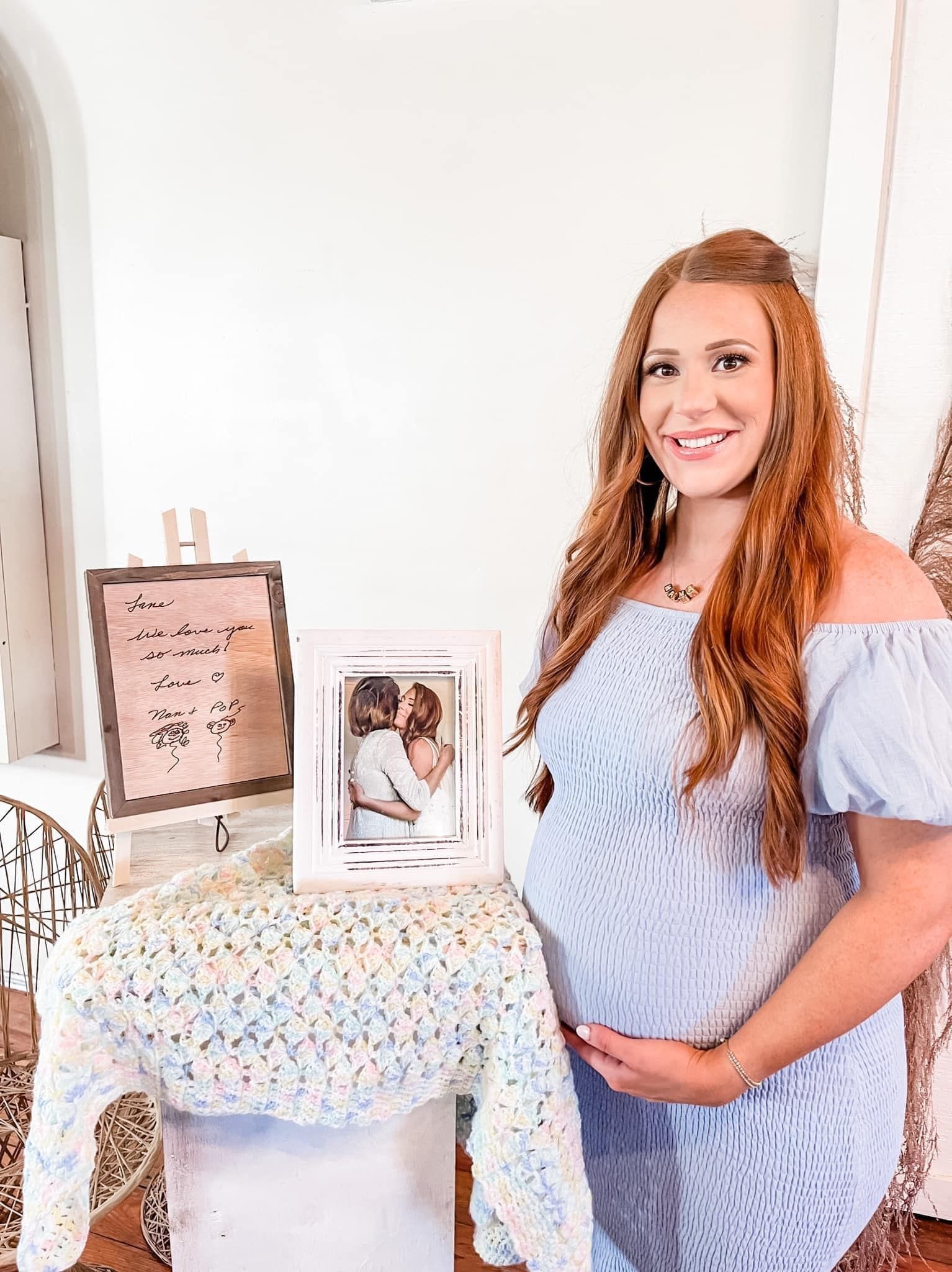A pregnant woman in a blue dress is standing in front of a table holding her belly.