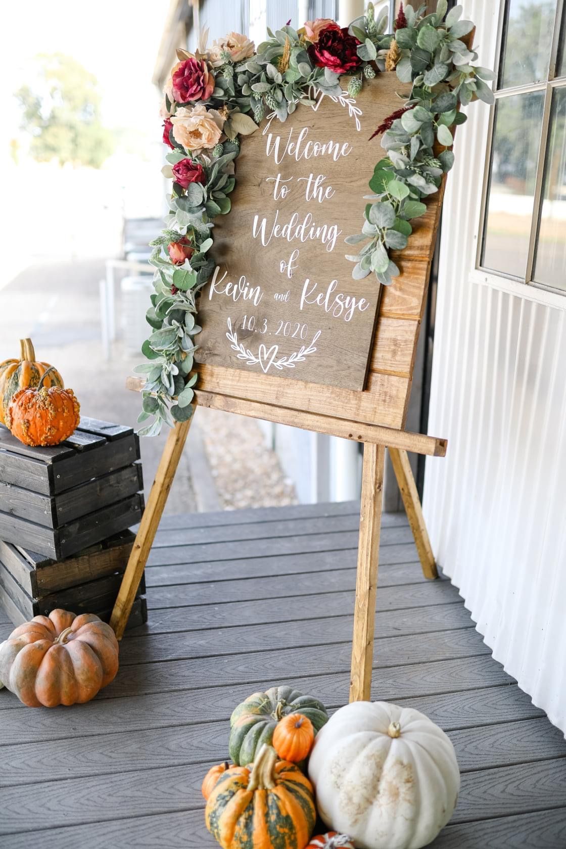 A welcome sign is sitting on a wooden easel on a porch surrounded by pumpkins.