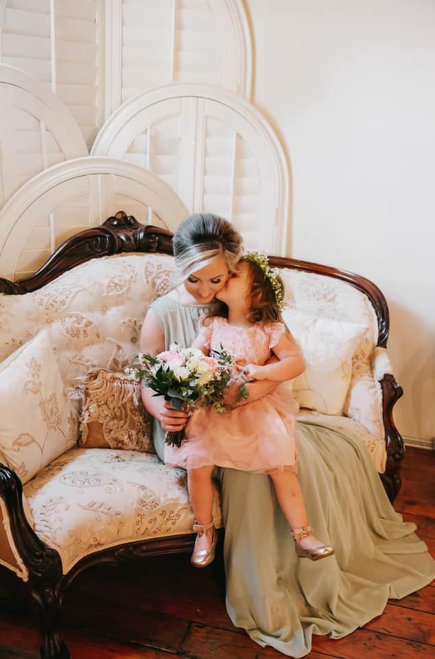 A woman is sitting on a couch holding a flower girl and kissing her on the cheek.