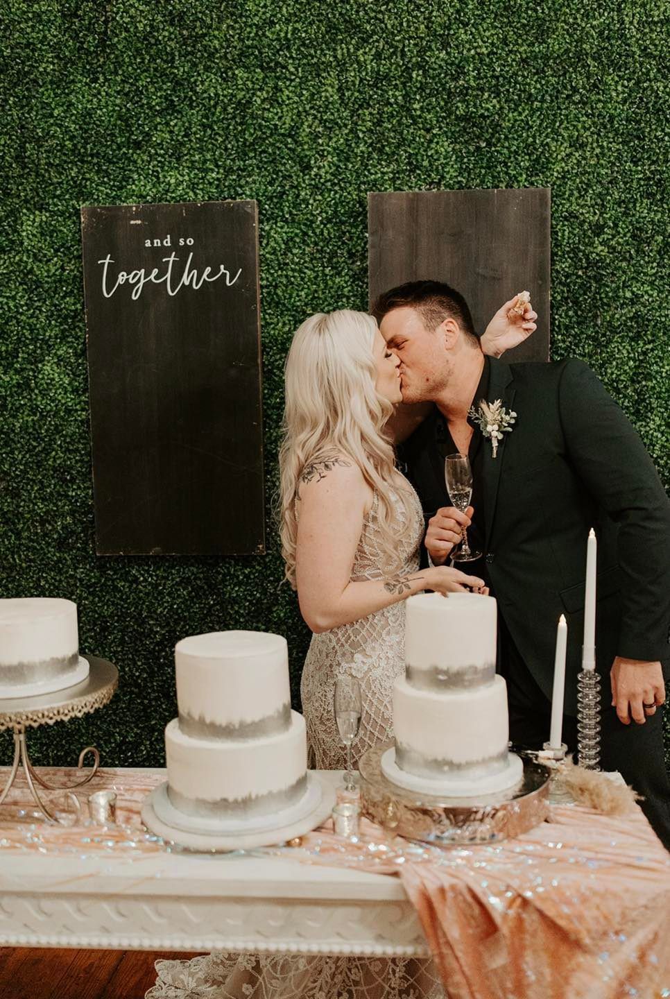 A bride and groom are kissing in front of a table with cakes.