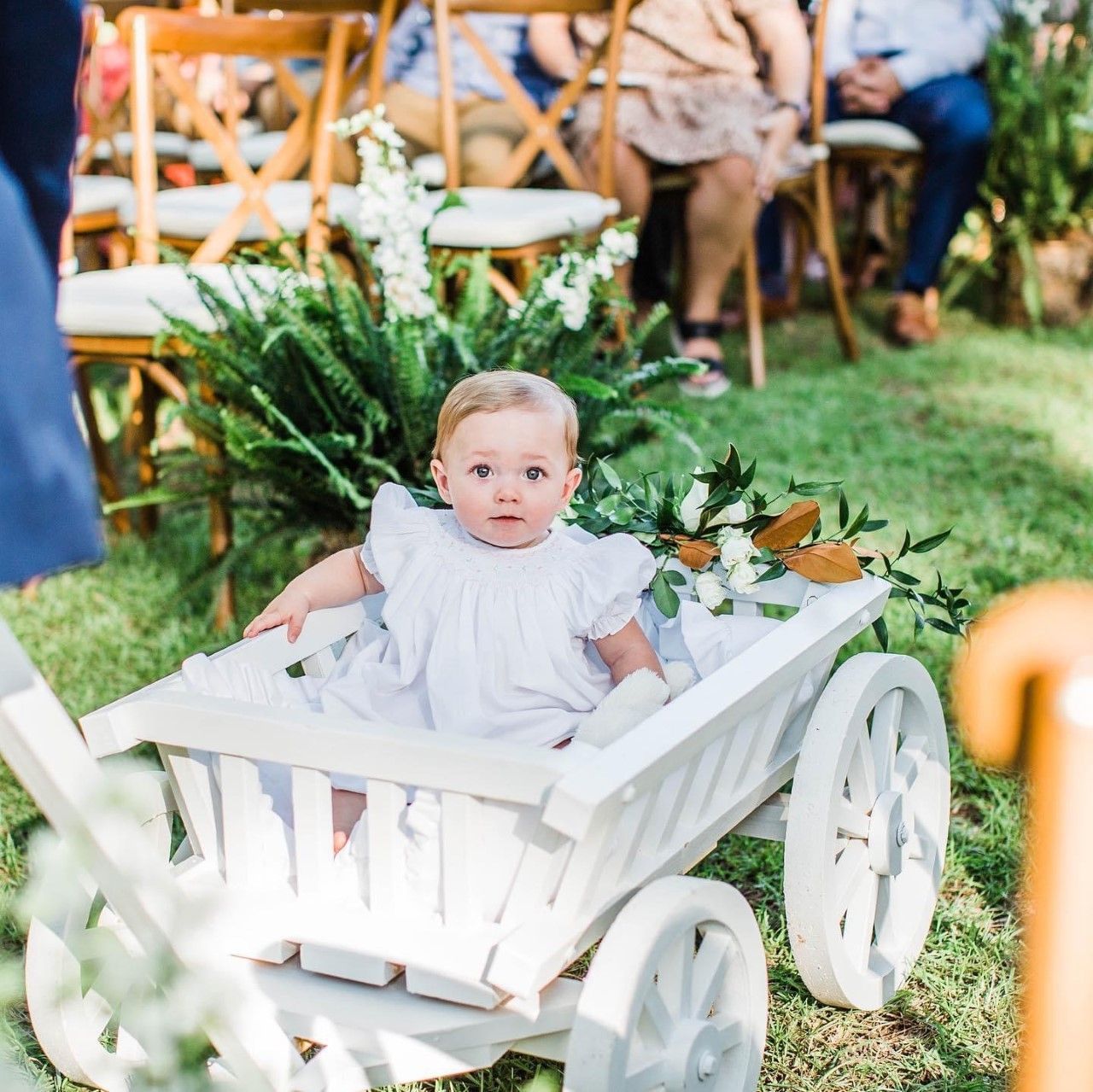 A baby in a white dress is sitting in a white wagon.