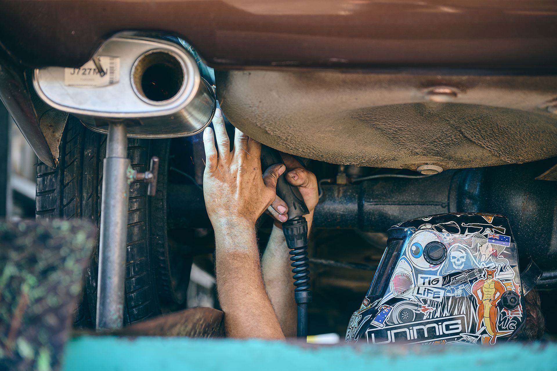 Mechanic working under a car, holding a tool near an exhaust pipe.