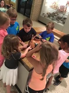 A group of children gathers around a display table in a museum to examine a small, brown, circular object.