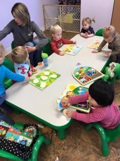 An adult supervisor sits with six children at a table, all engaging with various puzzles in a classroom setting.