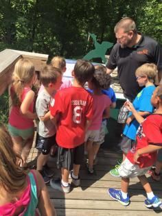 A group of children and an adult look at a display sign on an outdoor wooden deck surrounded by trees.